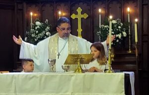 Fr. Gill saying the Eucharistic prayer with a boy on his right and a girl on his left, behind the altar.